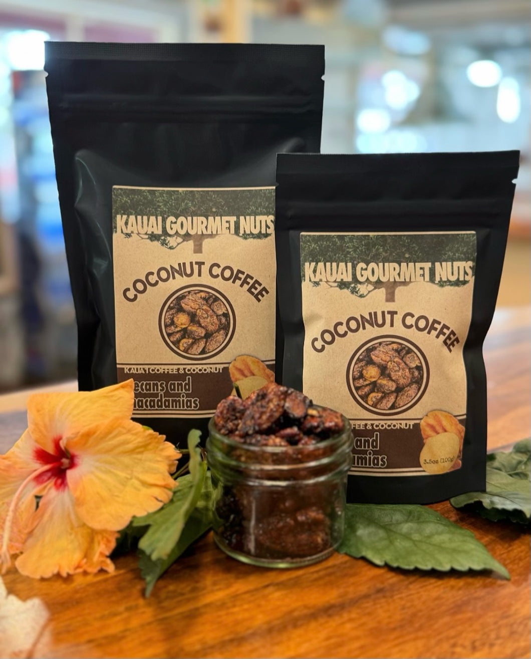 Two packages of Kauai Gourmet Nuts Coconut Coffee on a wooden surface with a flower and jar of coffee beans.